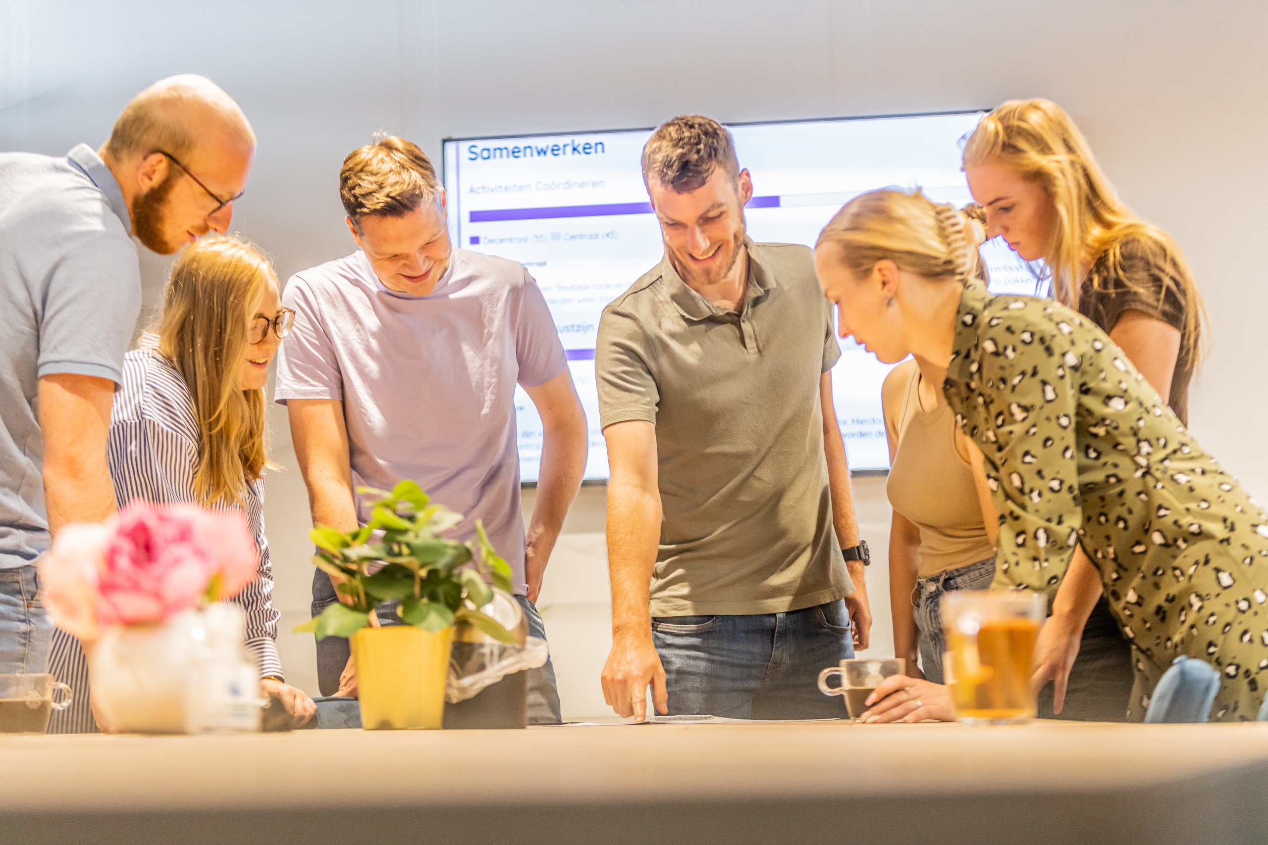 Team bespreekt een opdracht terwijl een trainer aanwijzingen geeft en een rapport over samenwerken zichtbaar is op de achtergrond.